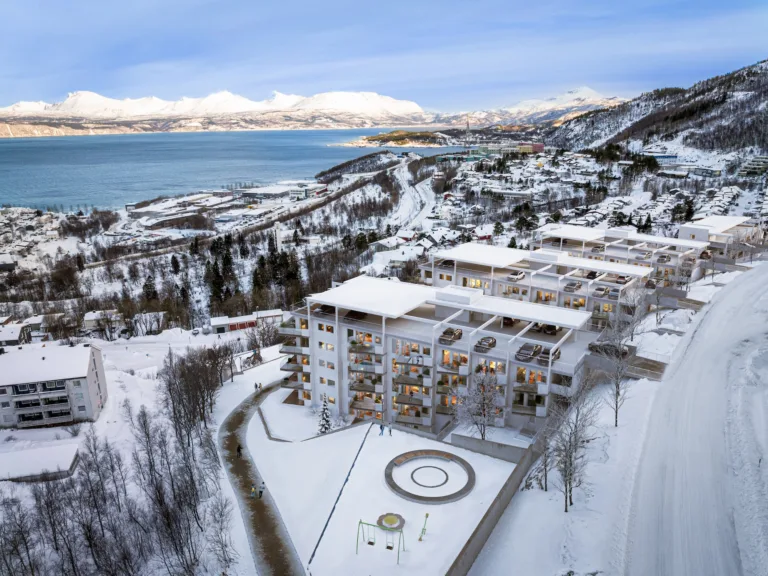 Flyfoto av et moderne leilighetskompleks omgitt av snø med fjell og vann i bakgrunnen.