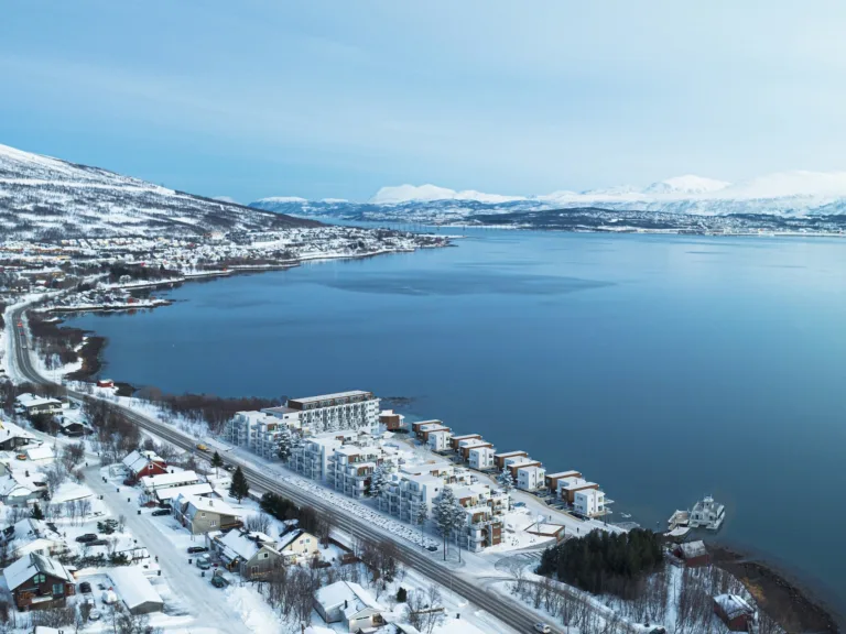 Flyfoto av en snødekt kystby med bygninger langs strandlinjen, et rolig, blått vann og fjell i bakgrunnen under en klar himmel.